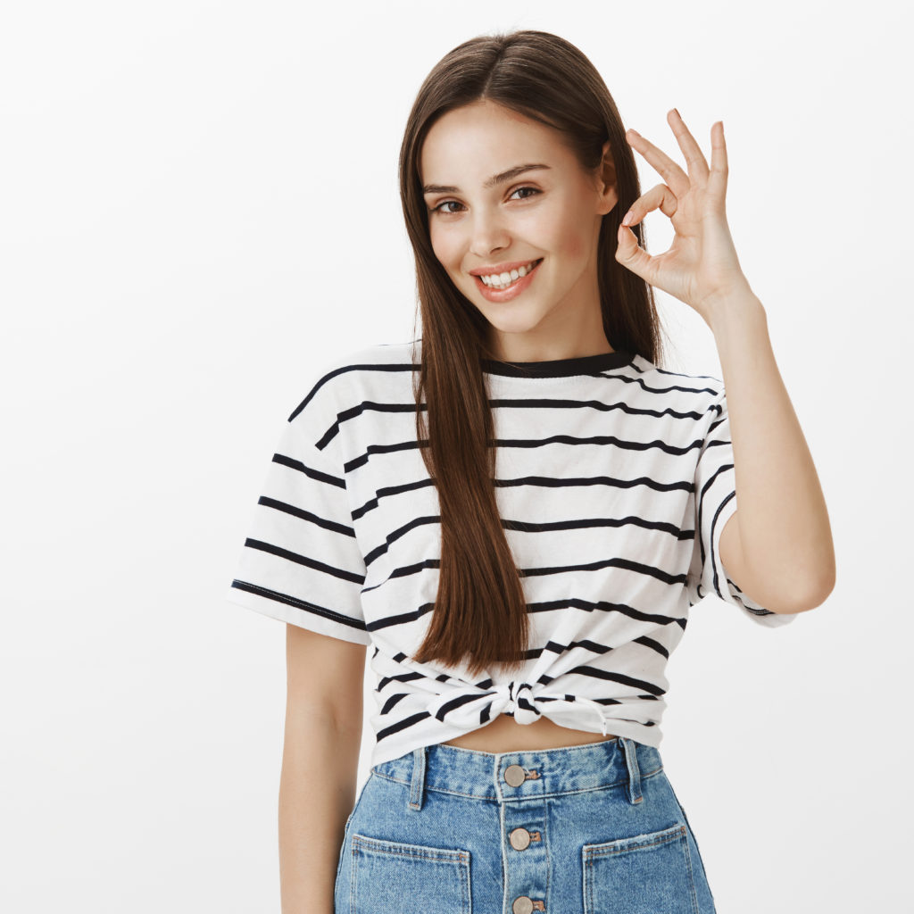 Girl attracts customers to her flower shop. Studio shot of positive confident european woman in striped t-shirt, raising hand, showing okay or great sign, giving approval and smiling broadly. Advertisement concept